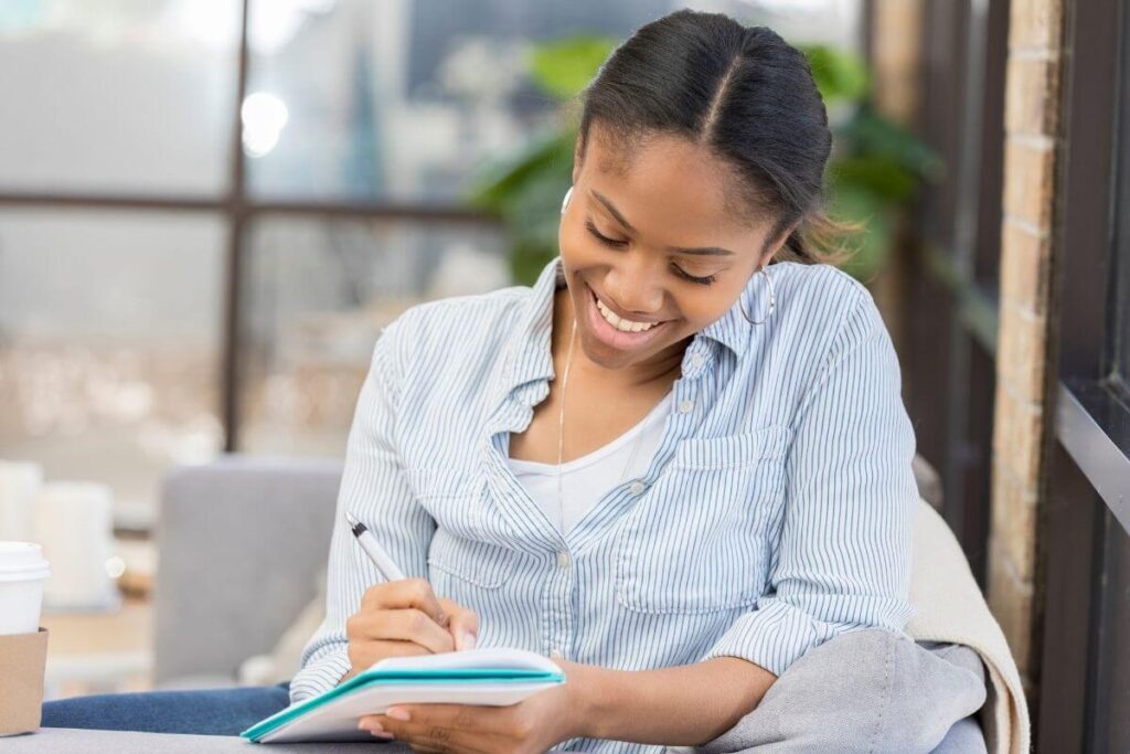 a happy young woman sits in a comfortable chair at the end of her work day writing in her business journal using journal prompts for entrepreneurs