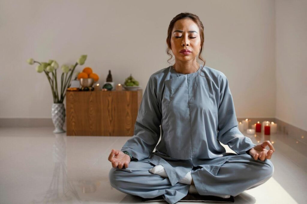 an asian woman sits in a meditation space in a yoga position with candles and other meditation room essentials around her as she peacefully meditates