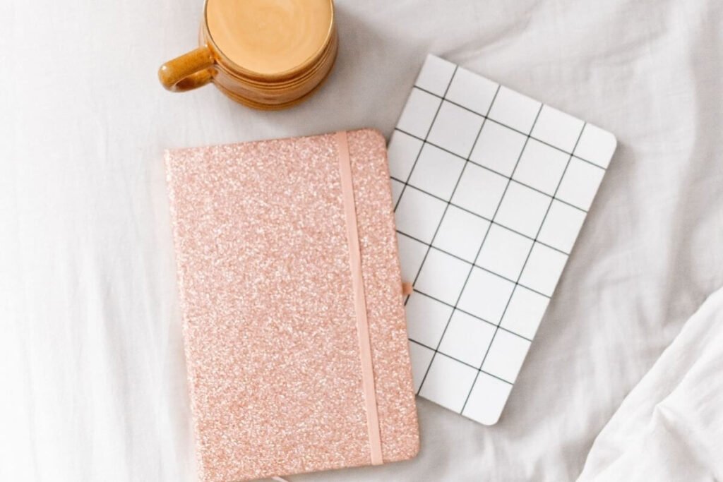 a top view of a glittery pink journal and a white and black journal sitting on a white tablecloth with a mug of frothy coffee as someone prepares to write about mindfulness journal prompts