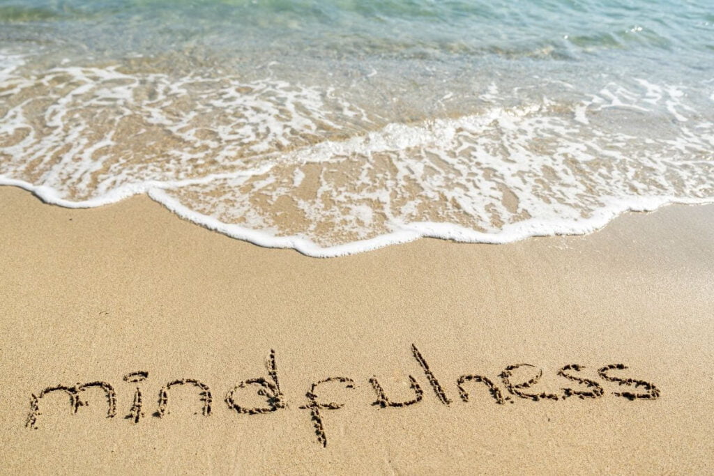 a calming close-up image of a beach shoreline with mindfulness written in the sand