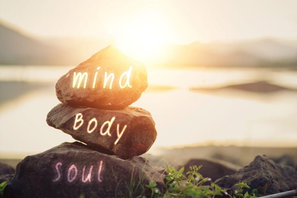 a close-up of three rocks that say mind body soul balancing on top of each other on a rocky beach against a blurred sunset