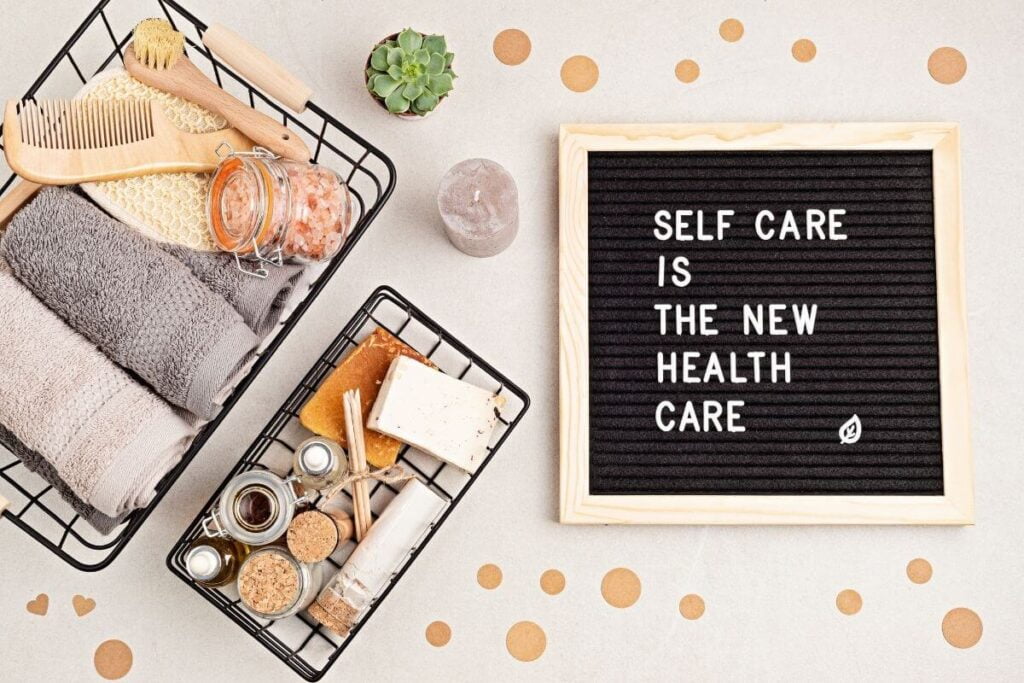 a sign that says self care is the new health care laying on a beige background with two baskets filled with self care items next to it