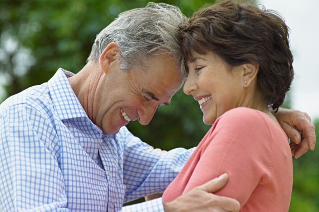 a middle-aged couple laughs together with their foreheads touching as they wander through a park with green trees