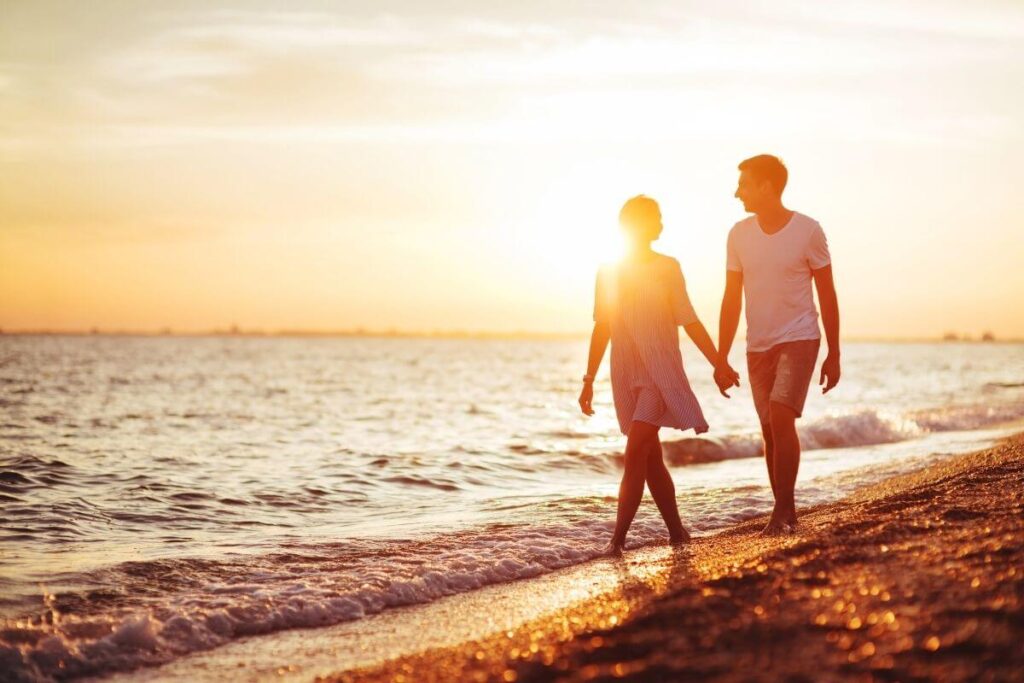 a couple walks along the seashore in the waves with the sun setting in the background