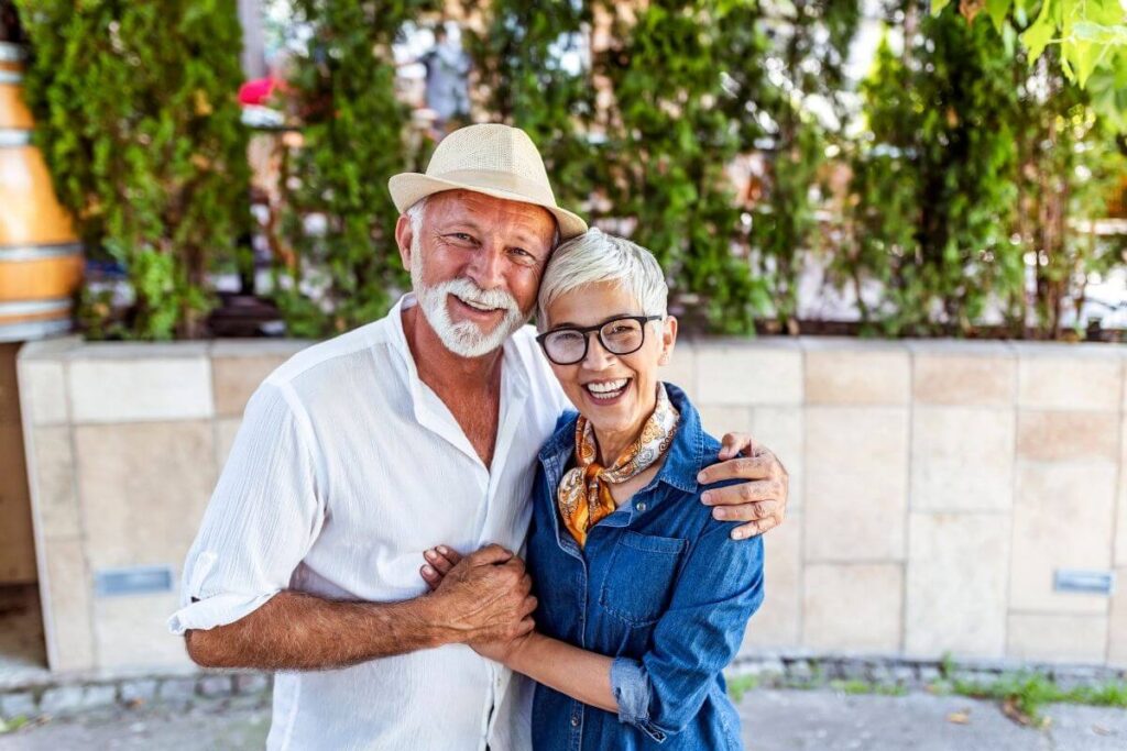 an elderly couple cheerfully embrace and smile for the camera in front of a concrete wall with greenery in the background