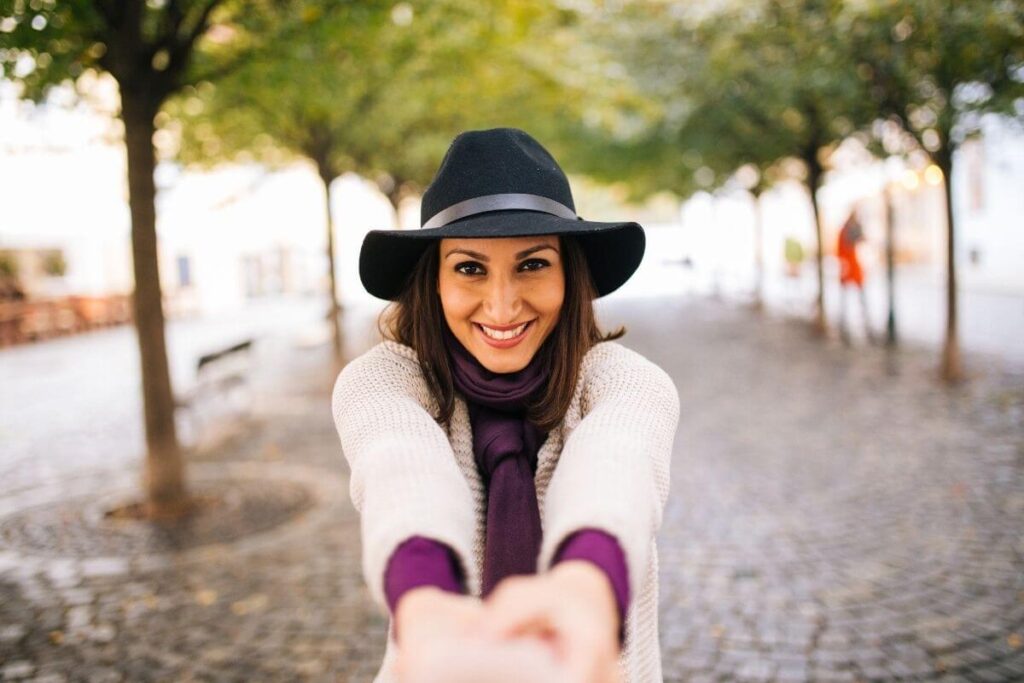 a middle aged woman wearing a black hat standing on a cobble stone path surrounded by trees proudly looks at the camera with her hands freely reaching out and shows that she has used her affirmations for letting to go to release negative energy