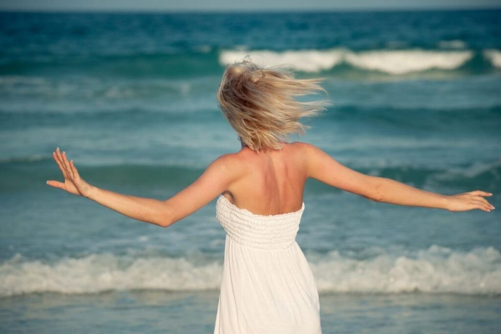 a blonde haired woman whimsically moves on the shore as the waves crash around her and she lets go of her troubles