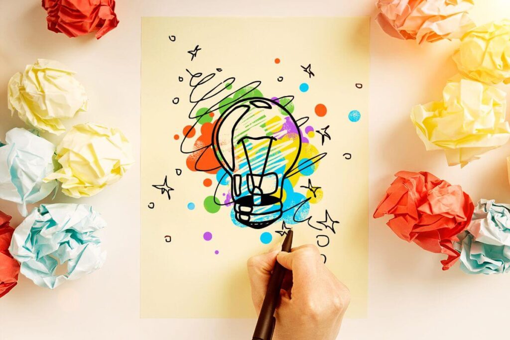 a top view of a hand drawing a colorful and creative lightbulb on yellow paper with balls of crunched up papers laying around like the artist needs affirmations for creativity because she has a creative block