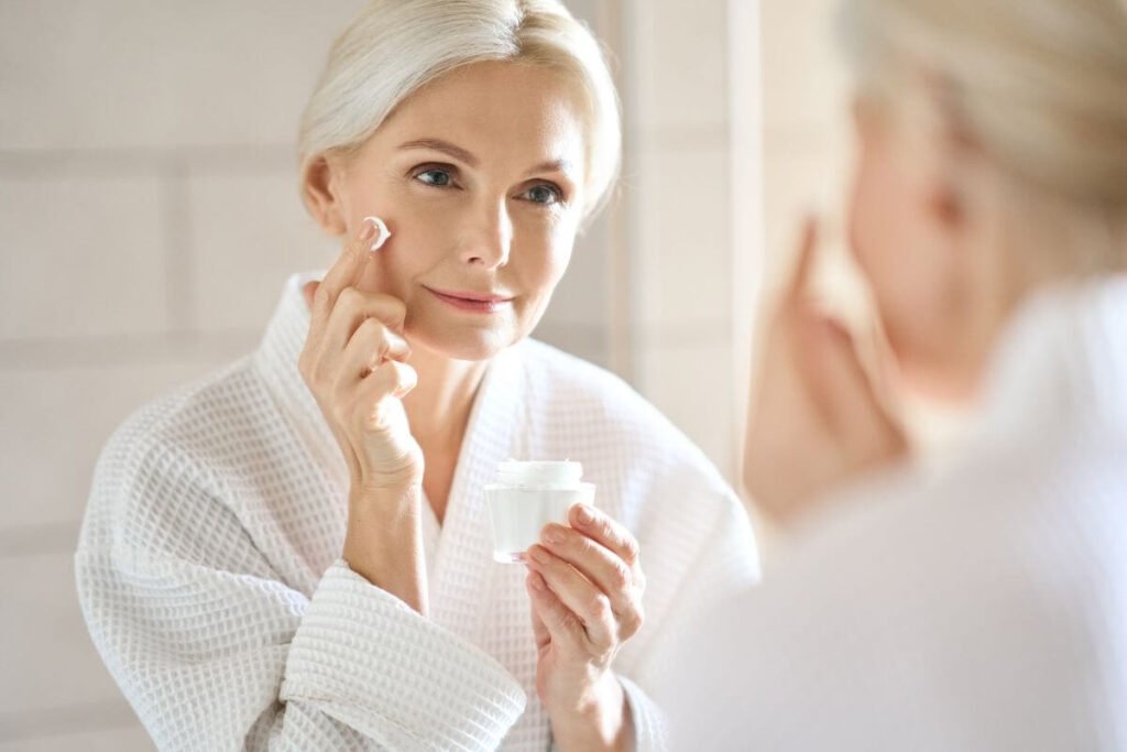 a middle aged blonde woman in a white bathrobe looks into the mirror as she practices self love with her skin care