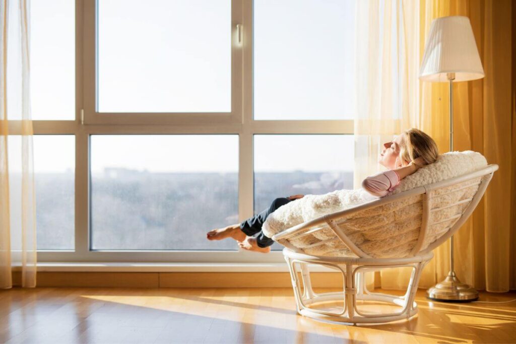 a young woman lounges in a circle chair in front of a large window with the sun warming her as she relaxes and shows signs of good personal growth