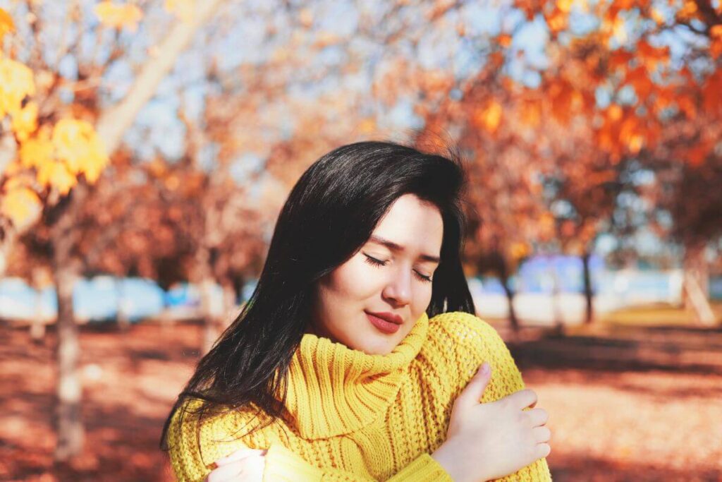 a young woman with long dark hair in a beautiful yellow sweater gives herself a hug for self care outside in a park in the fall