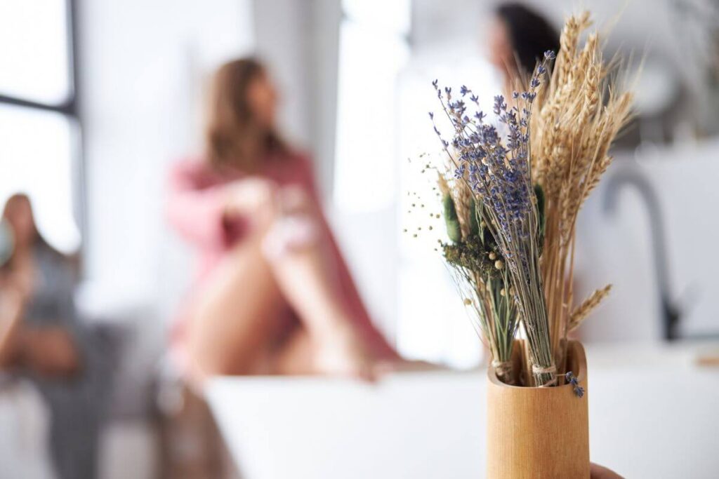 two out of focus women are sitting in the background of a photo relaxing in an all white environment and a wooden vase sits in the foreground with some dried flowers in it