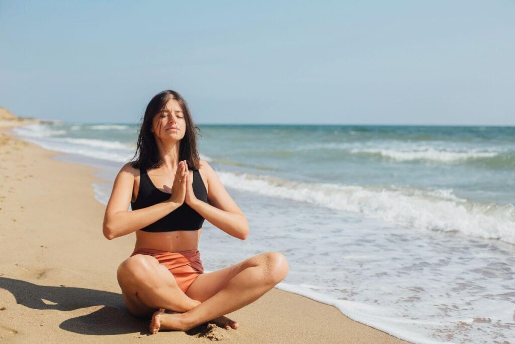 a young women with brown hair wearing shorts and a tank top does yoga along the shoreline with the ocean in the background as she demonstrates unusual self care ideas