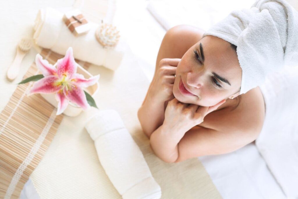 a young woman ponders what is wellness and why is it important as she relaxes on a massage table with her chin resting on her hands and a pink lotus flower in front of her