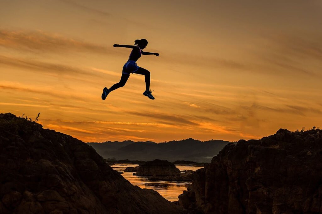 an athletic young woman courageously leaps from one hill to another over a body of water