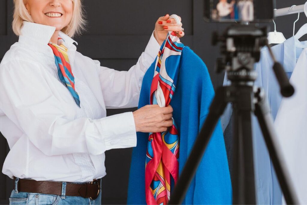 a middle aged blonde woman holds up a blue jacket with a bright colored scarf as she films a segment for her beauty blog for women over 50