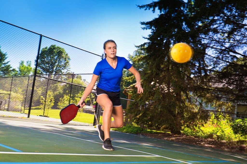 a middle aged woman in a blue tee shirt and black sorts plays pickle ball on an outside court