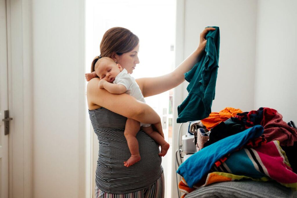 a young overwhelmed woman holds a baby and tries to fold laundry with one hand as she tries to use healthy coping skills