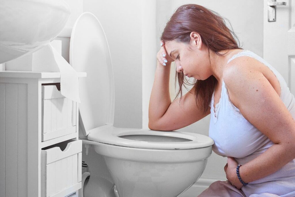 a young woman sits on the bathroom floor next to a toilet holding her stomach because she is struggling with stress and overwhelm and its impacting her physical health