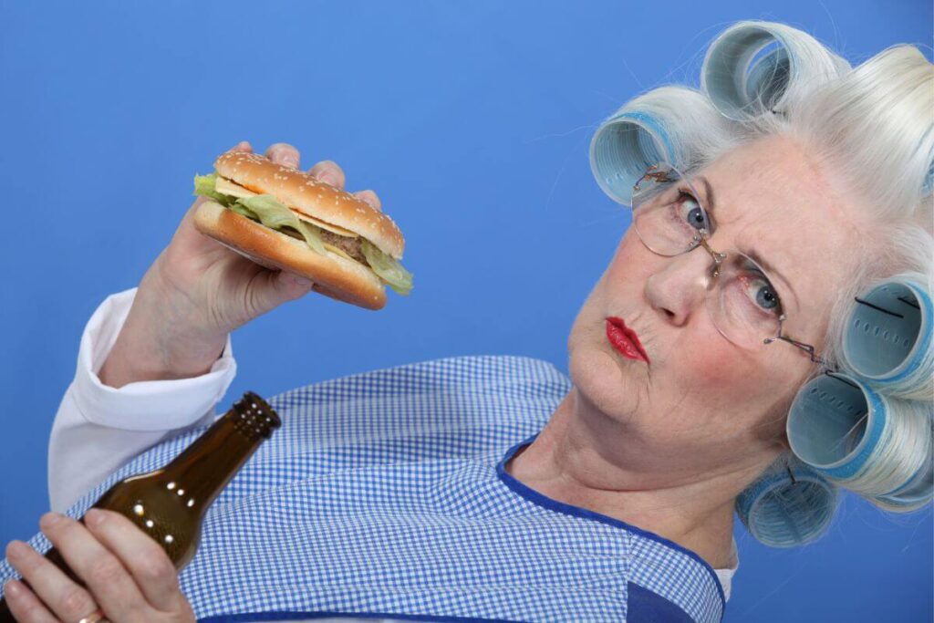 an older woman with her gray hair in big curlers eats a sandwich and holds a beer as she confidently stares at the camera with a sassy attitude