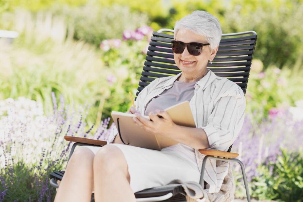 an older very happy woman sits in a lawn chair next to a purple flower garden and uses wellness journal ideas and prompts for her daily journaling practice