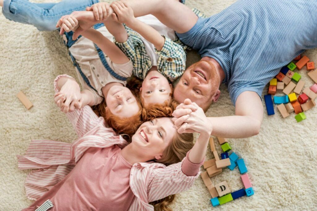 a young family of four lay in the floor with their heads together hold hands up in the air enjoying a happy life together