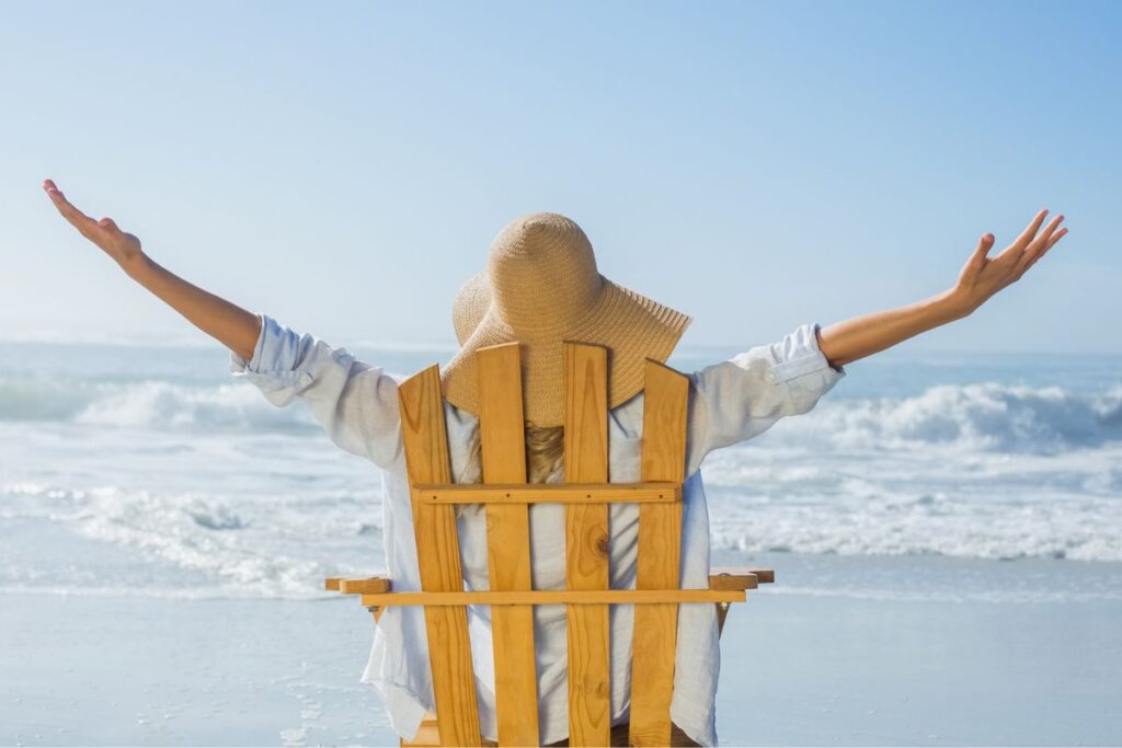 a woman stretches her arms to the sky in happiness because she has learned how to become unbothered in life sits on the beach in a wooden chair facing the waves