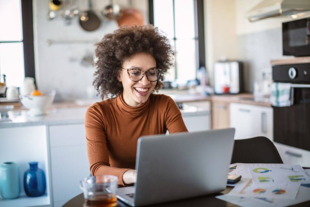 a happy woman works on her laptop at a table in her kitchen