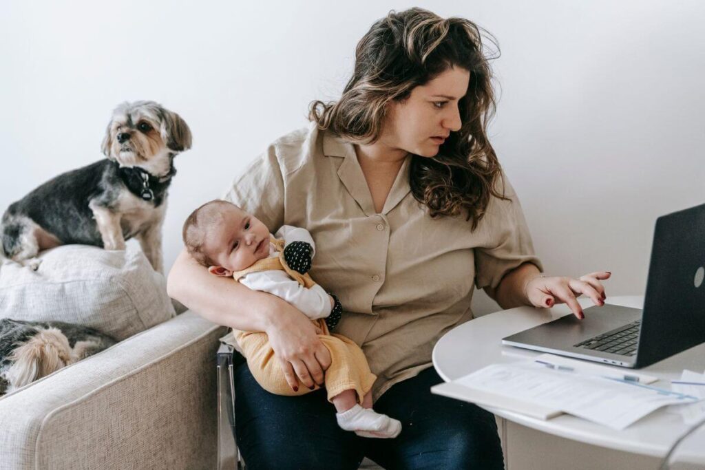 a busy mom holds a baby and has a dog peeking over her shoulder as she does not maintain a healthy lifestyle while working from home