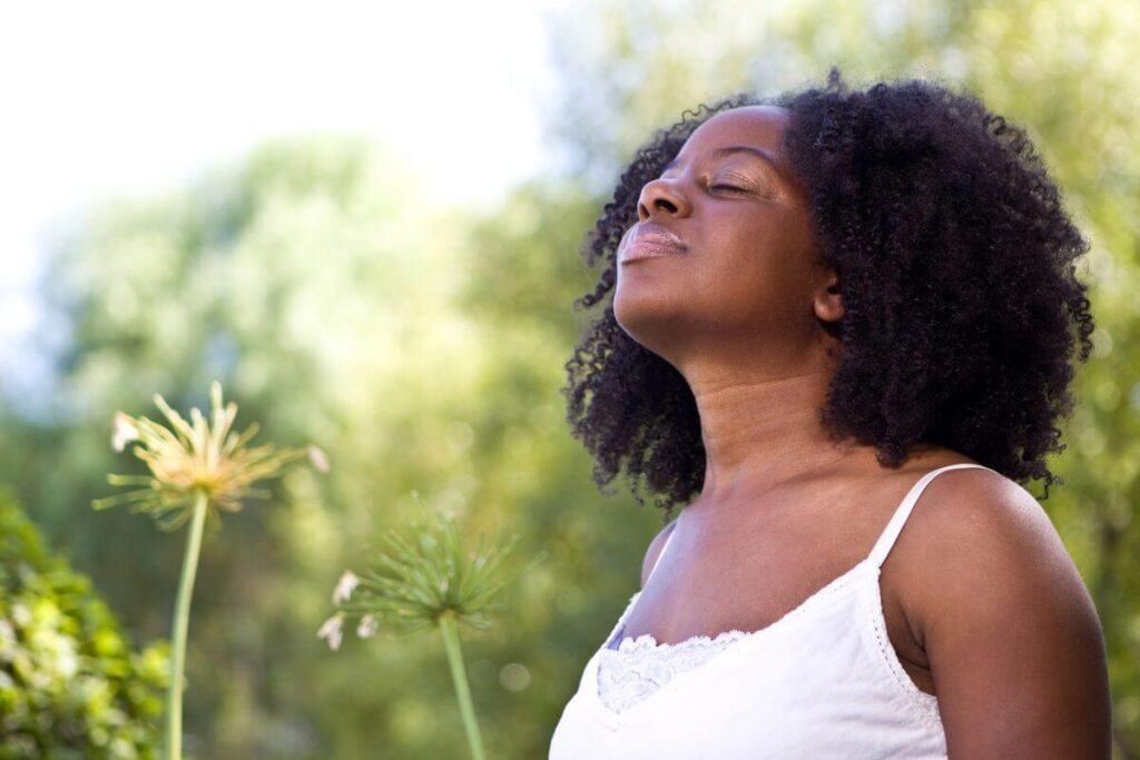 a dark skinned young woman with her eyes closes and a smile on her face breathes in the fresh air in a park