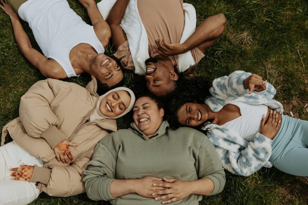 5 women of varying sizes shapes and styles lay on the ground together laughing and enjoying life in their individual bodies