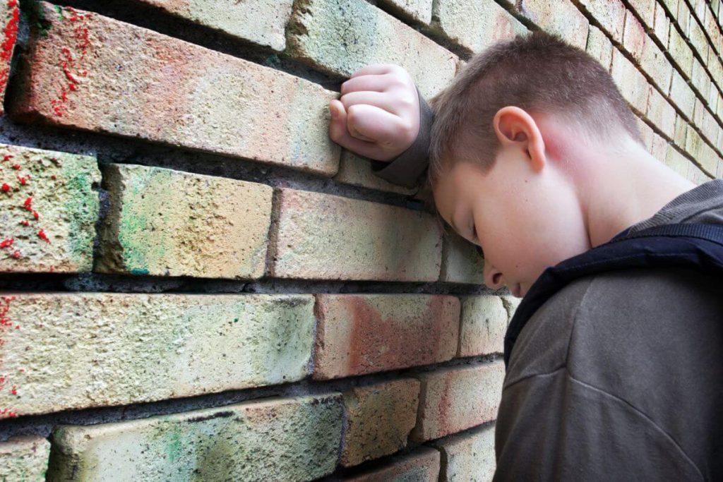 a young boy is excluded on the playground and stands alone with his head against a brick wall