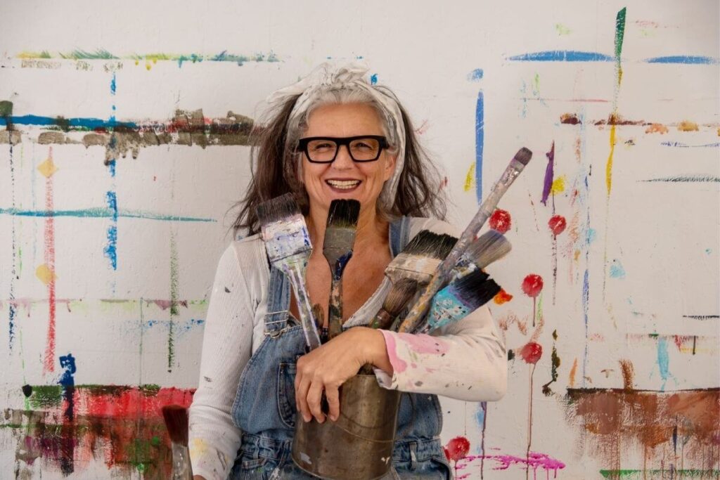 a sassy and confident but messy middle aged female artist holds used paint brushes in front of a colorful canvas