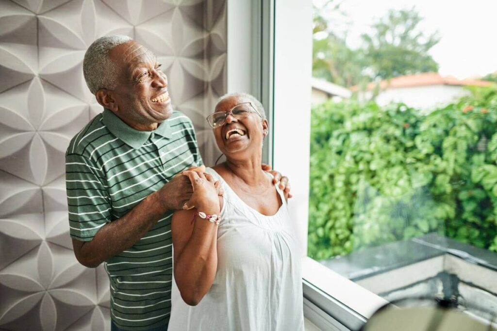 an elderly black couple stand in front of a large window laughing together as they live consciously