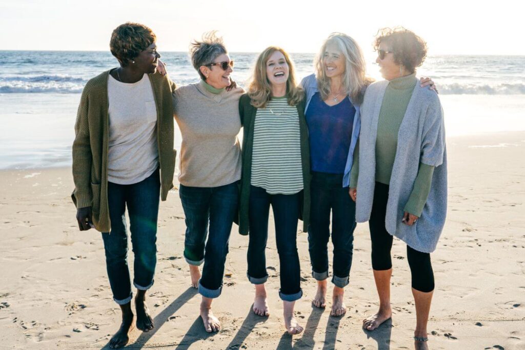 five middle aged women gather together on the beach in bare feet and jeans laughing and hugging