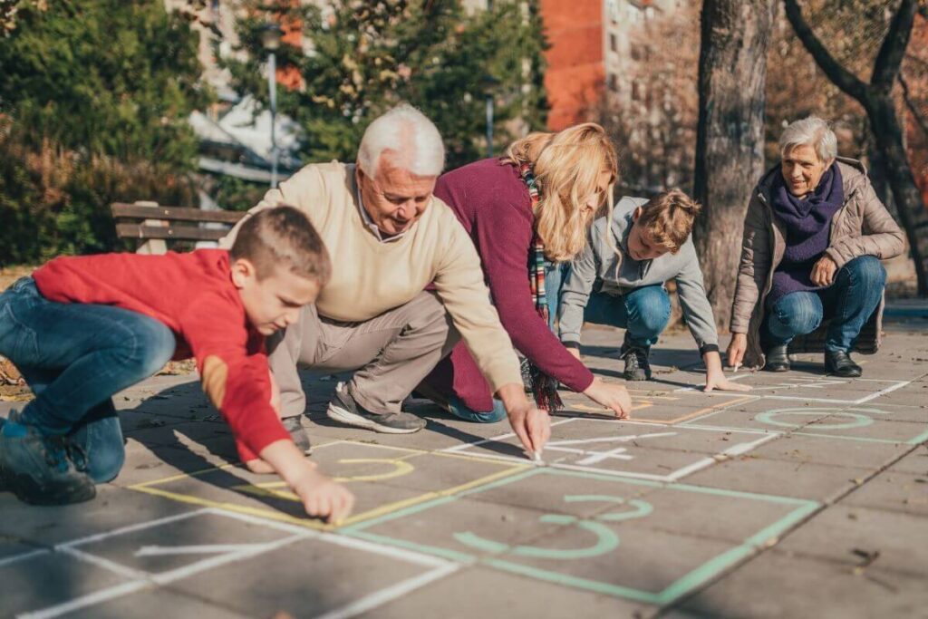 five members of a multigenerational family who realize the benefits of living with elderly parents create a hopscotch game together with sidewalk chalk