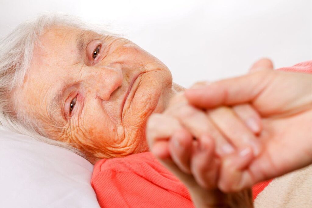 an elderly woman lays in bed at a nursing home holding hands with a loving daughter who visits often