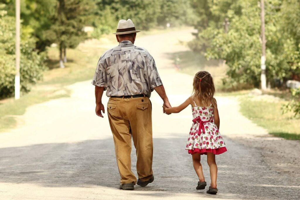 elderly grandfather walks down a dirt road with his young granddaughter