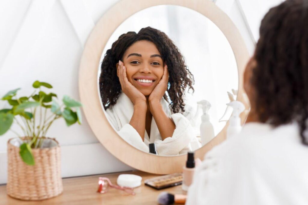 a beautiful black woman relaxes during her self care routine and looks happily and proudly at herself in the mirror