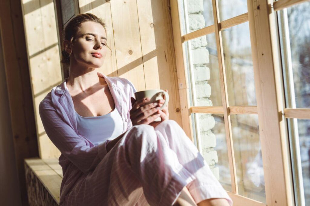 a women enjoys the morning sunshine as she relaxes on a window seat in her home with a cup of coffee