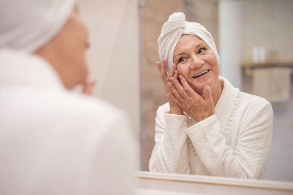 a middle aged woman getting ready for the day by washing her face at the bathroom mirror with a bright smile on her face