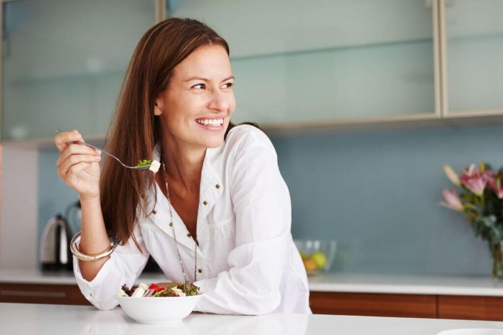 a happy women stands at the island in her minimalist kitchen space and eats a bowl of simple salad