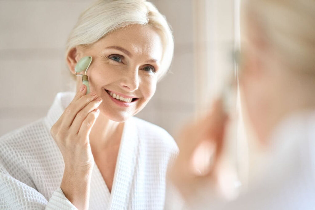 a middle aged woman blonde woman practices self care she uses a jade roller on her face and looks in the mirror