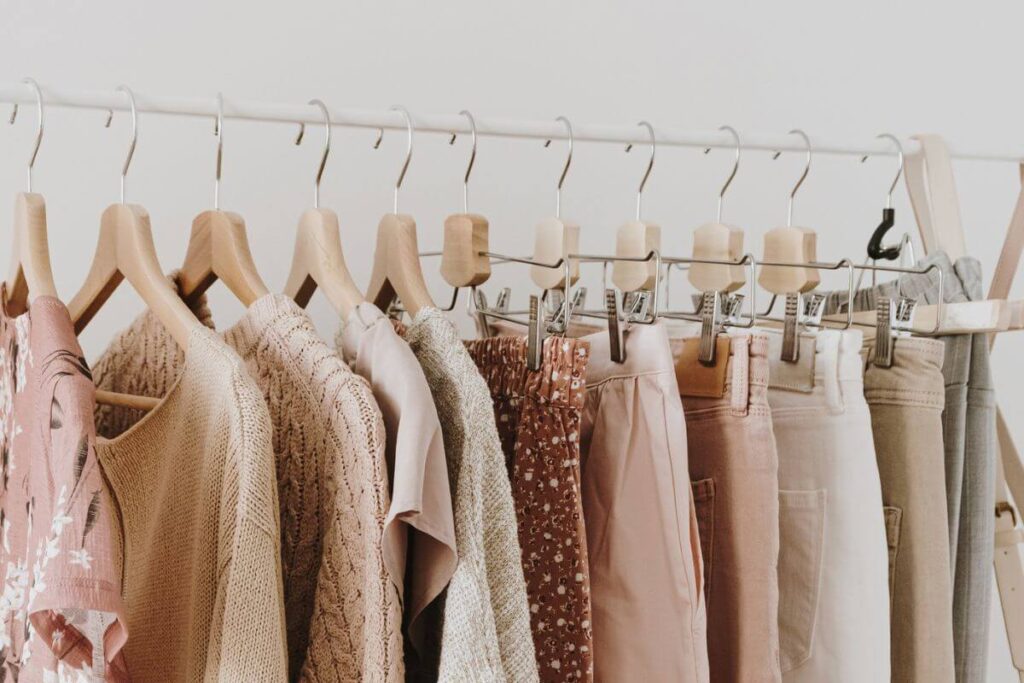 minimalistic capsule wardrobe of sweaters, pants, and blouses in shades of pinks and neutrals hang on a clothes rack