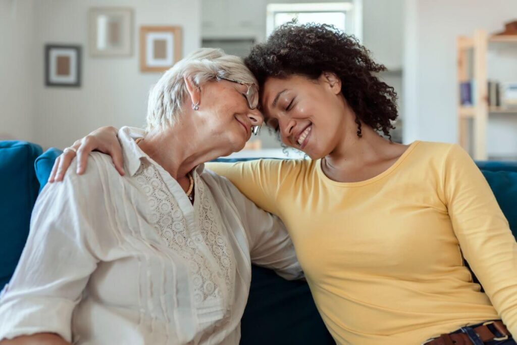 an adult women and her elderly mother sit on a sofa and hug after having an honest conversation about expectations