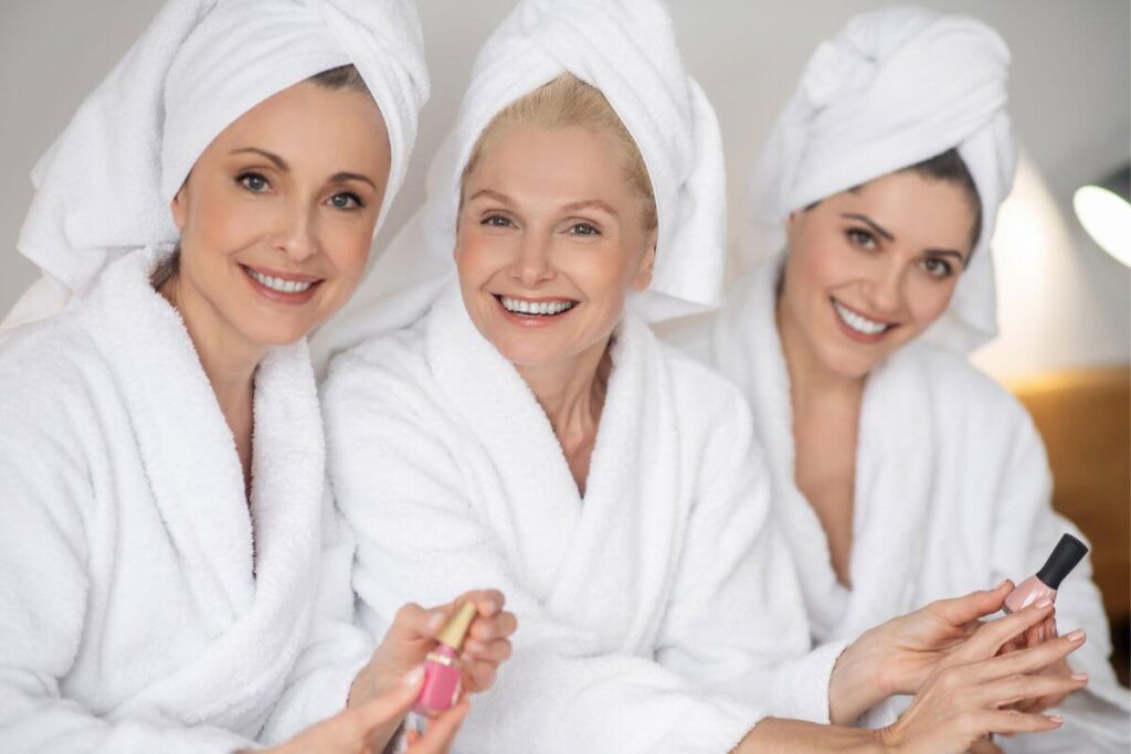 three women over 50 sit together in robes with towels wrapped around their hair waiting for pedicures and manicures at a spa