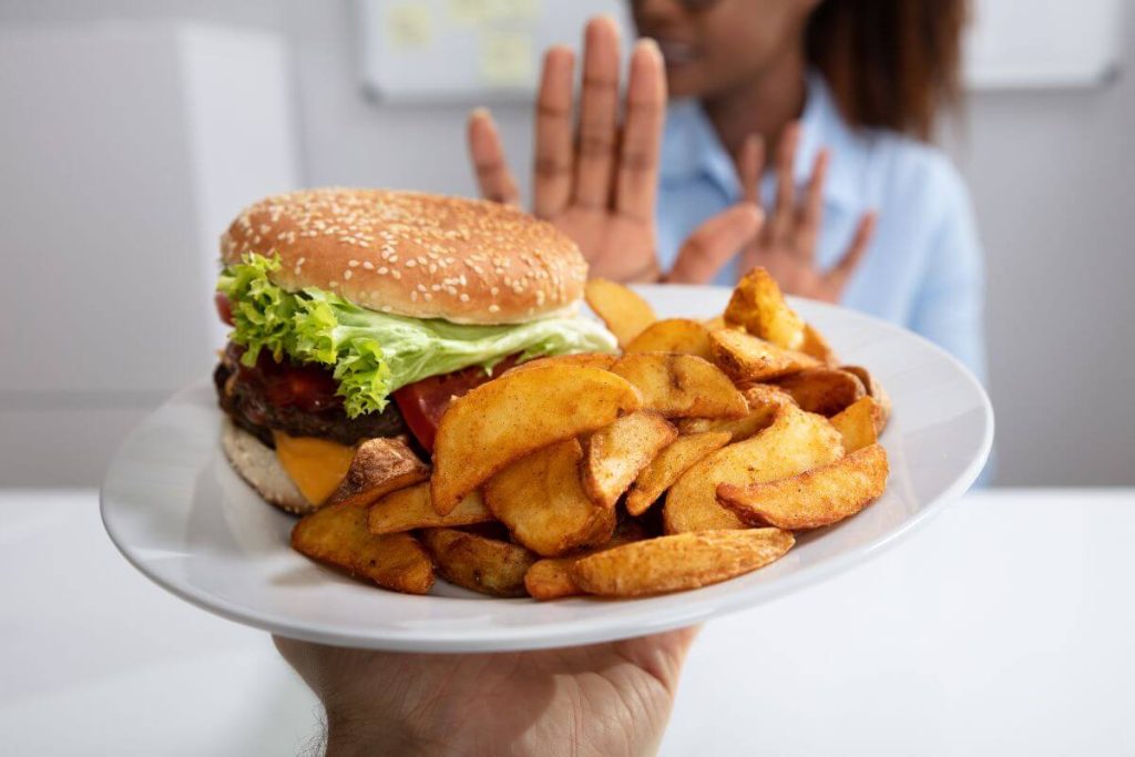 a woman in the background holds up a hand to say no thank you to a plate of french fries and a large burger because she knows it's not healthy for her as she ages