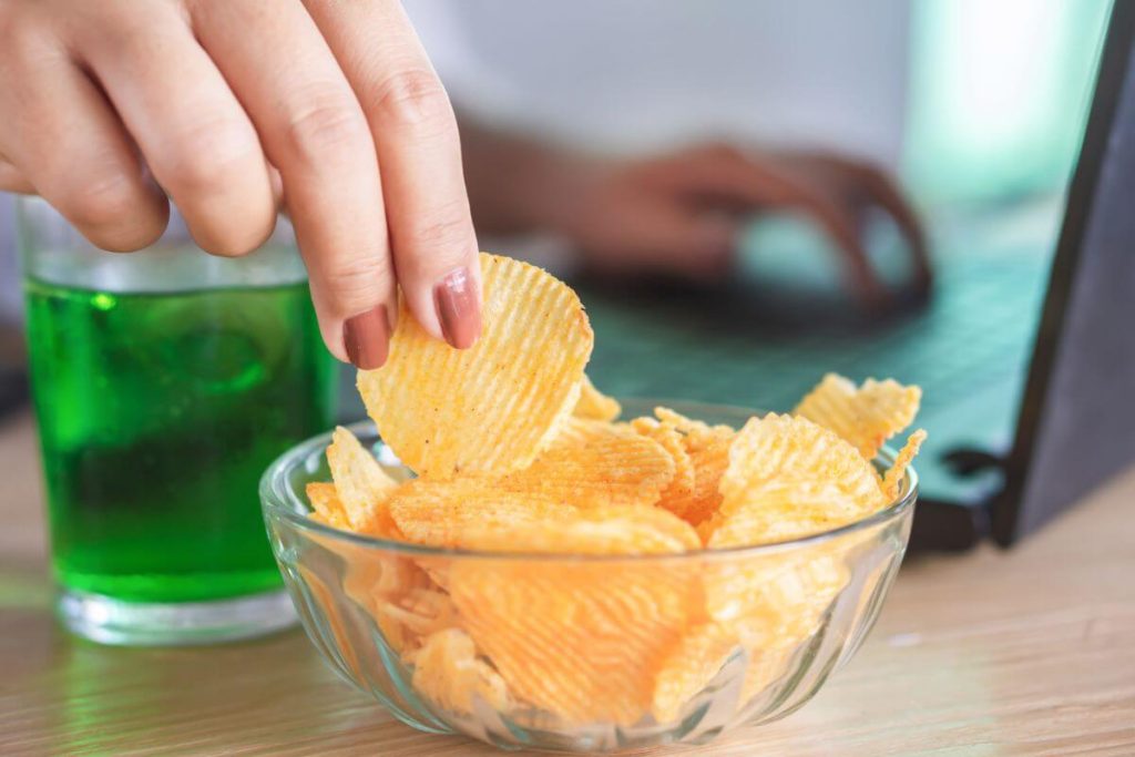 a close up of a female picking up a potato chip from a bowl and a sugary drink sitting next to the bowl