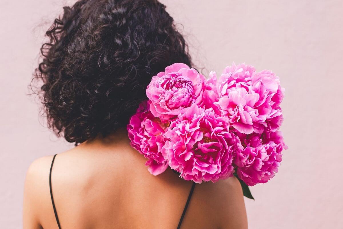 close up of the back of a woman holding a bouquet of beautiful pink flowers over her shoulder that she bought to be kind to herself