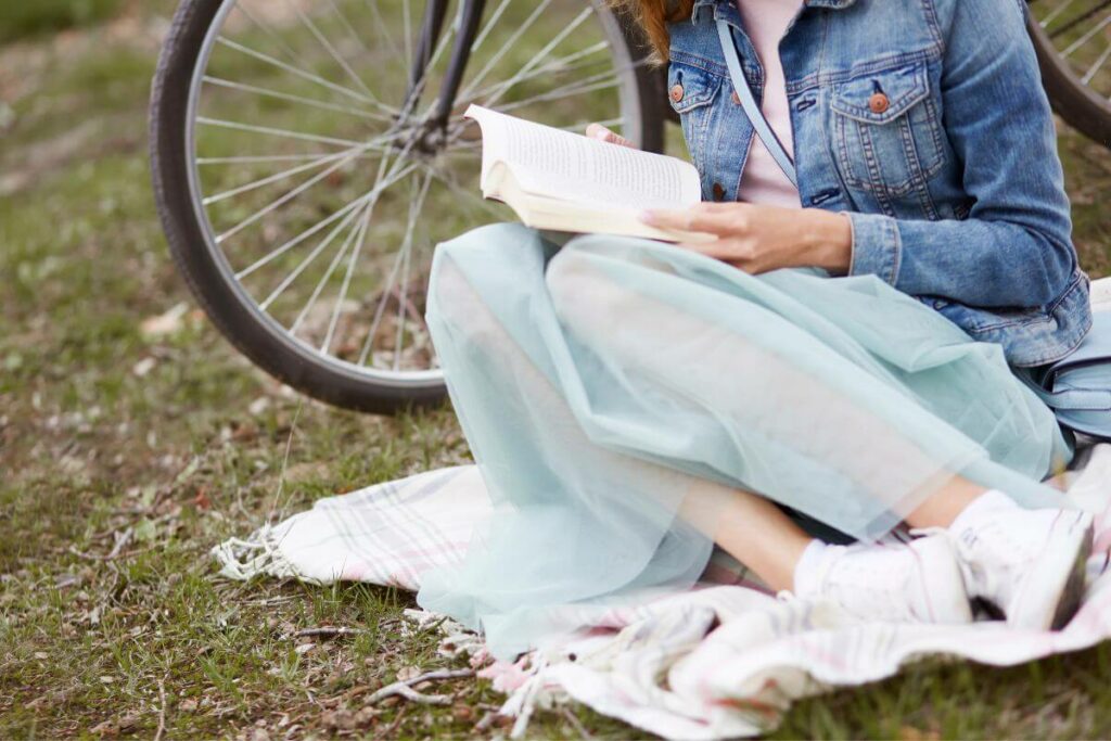 a women sits next to a bicycle on a small blanket to relax and read outside in a park